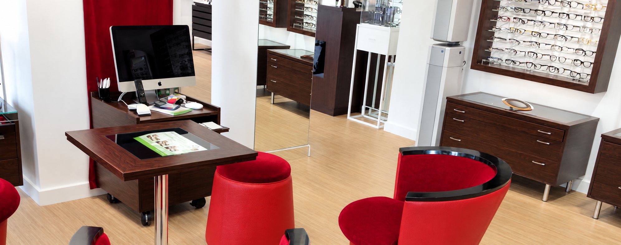 Intérieur d'un magasin d'optique moderne avec fauteuils rouges, mobilier en bois sombre, ordinateur et présentoir mural de lunettes.