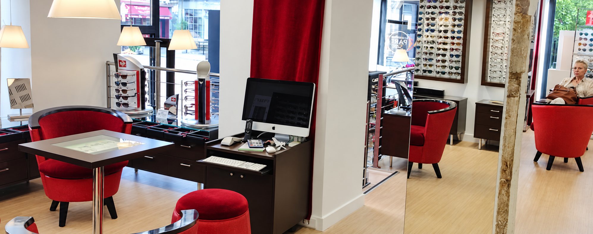 Intérieur lumineux d'un magasin d'optique avec des fauteuils rouges, un poste informatique et des murs de lunettes de soleil exposées.