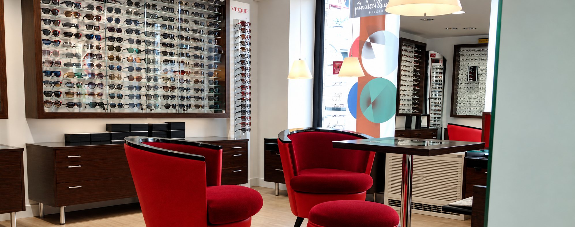 Intérieur lumineux d'un magasin d'optique avec des fauteuils rouges autour d'une table et des murs couverts de lunettes de soleil exposées.