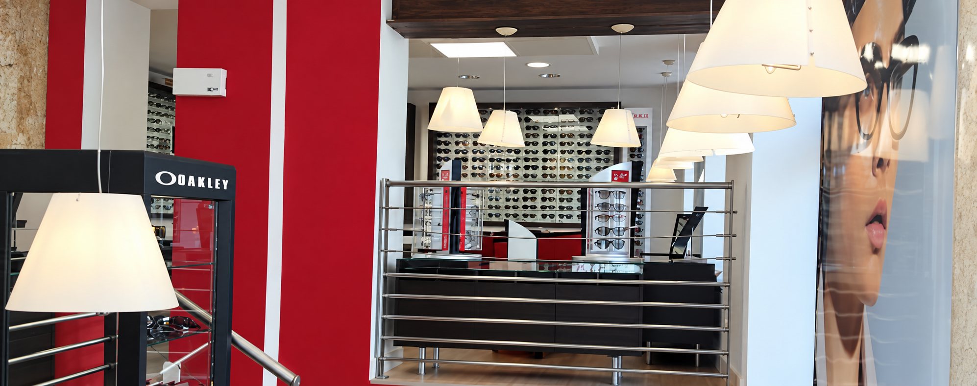 Intérieur d'un magasin d'optique aux murs rouges et blancs, avec des suspensions blanches, une vitrine Oakley, un comptoir et un mur de lunettes de soleil.
