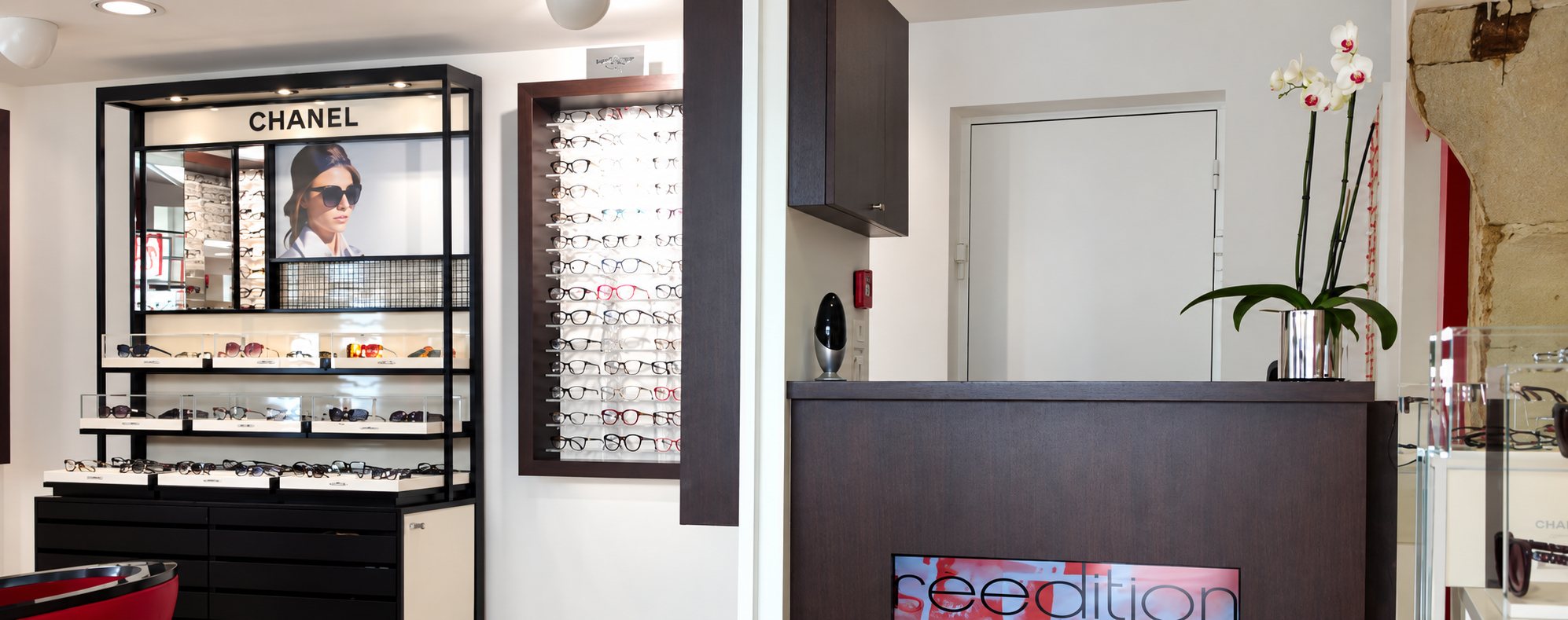 Intérieur d'un magasin d'optique avec un présentoir Chanel, un mur de lunettes et un comptoir en bois sombre orné d'une orchidée.