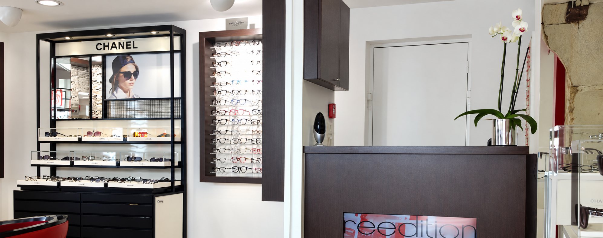 Intérieur d'un magasin d'optique avec un présentoir Chanel, un mur de lunettes et un comptoir orné d'une orchidée blanche.