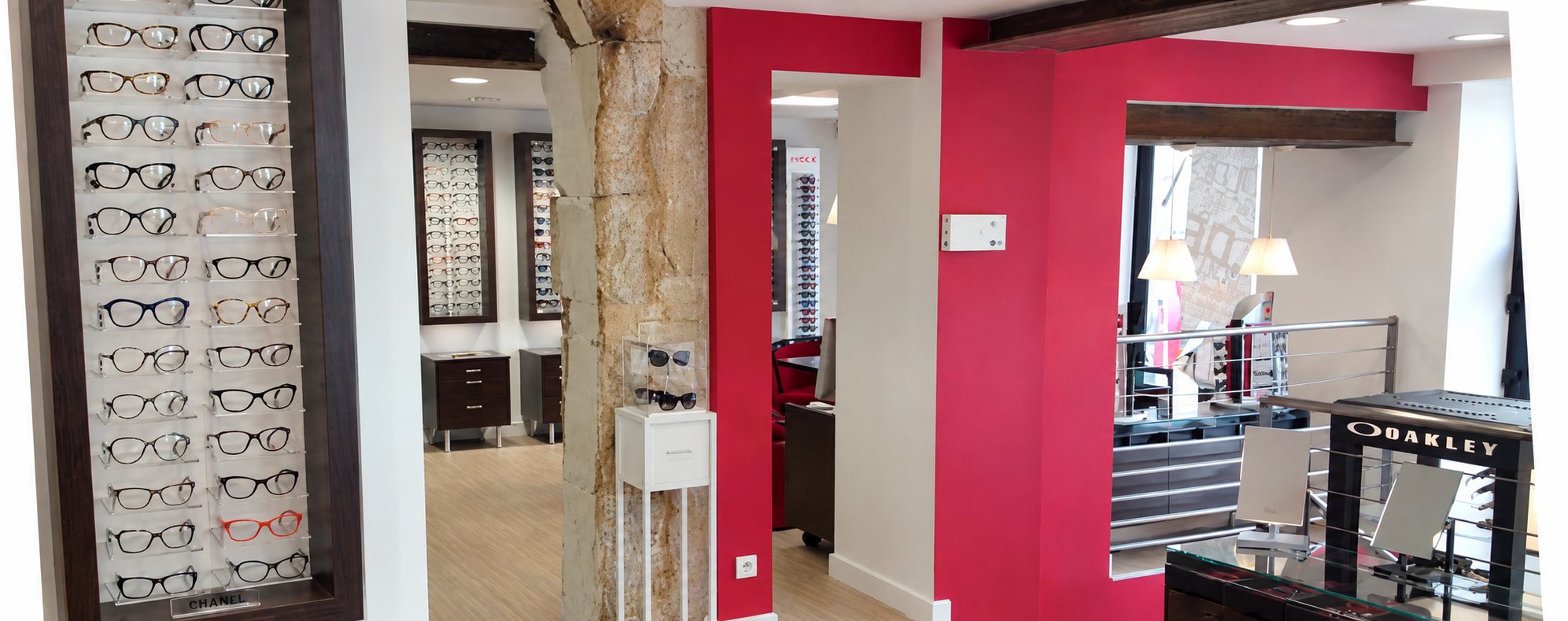 Intérieur lumineux d'un magasin d'optique aux murs rouges et blancs, avec des présentoirs muraux de lunettes de vue et un meuble Oakley.