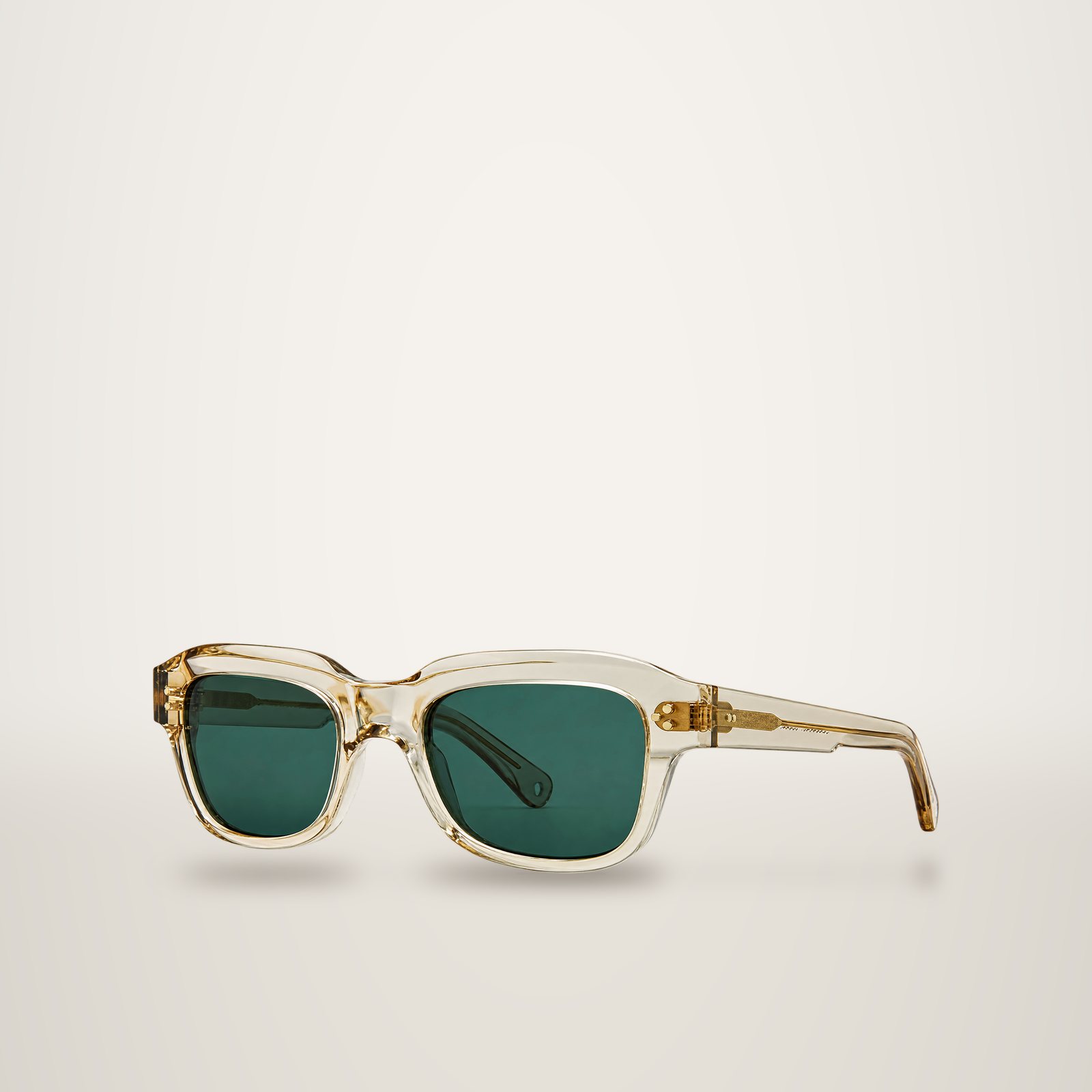 Une paire de lunettes de soleil à monture rectangulaire en acétate transparent beige, avec des verres vert foncé et des charnières dorées, photographiée sur fond blanc.