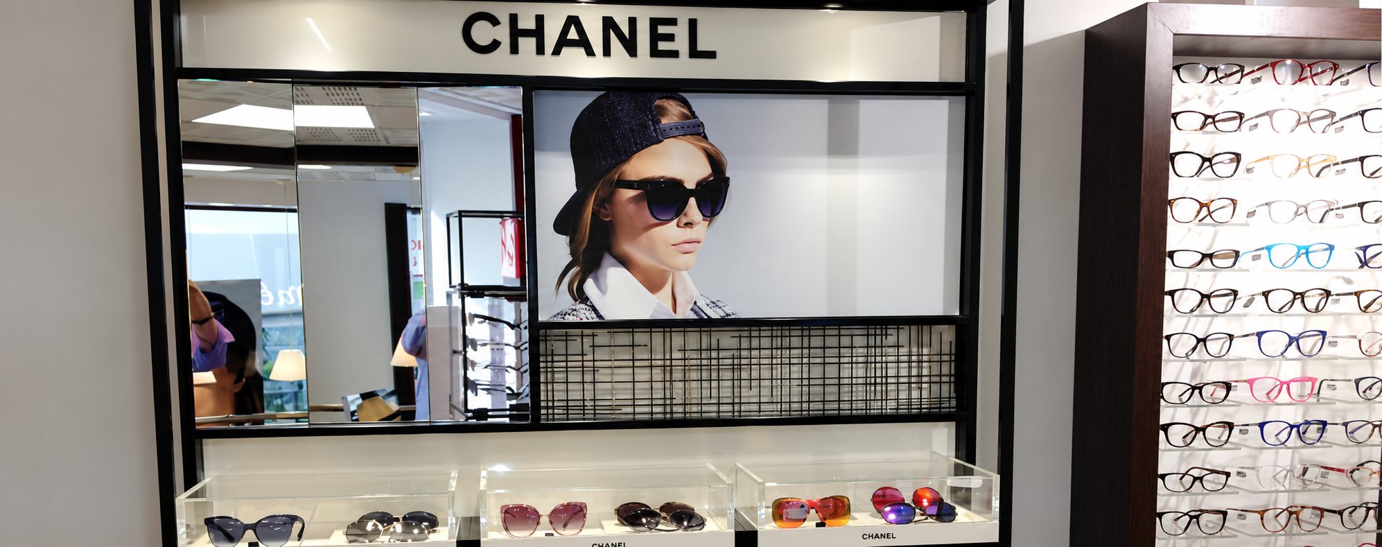 Un présentoir Chanel dans un magasin d'optique avec une affiche de mannequin en lunettes de soleil, des vitrines de montures colorées et un mur de lunettes de vue à droite.
